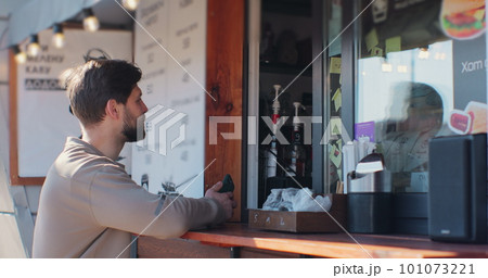Young man is buying takeaway coffe in coffee-shop and paying with smartphone making contactless payment. Modern technology and banking concept. 101073221
