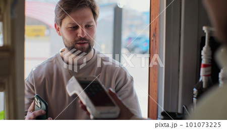 Young man is buying takeaway coffe in coffee-shop and paying with smartphone making contactless payment. Modern technology and banking concept. Young man is buying takeaway coffe in coffee-shop and paying with smartphone making contactless payment. Modern technology and banking concept. 101073225