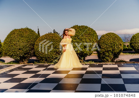 Woman yellow dress chess. A beautiful woman in a long puffy yellow dress poses on a chessboard in the park. Woman yellow dress chess. A beautiful woman in a long puffy yellow dress poses on a chessboard in the park. 101074848