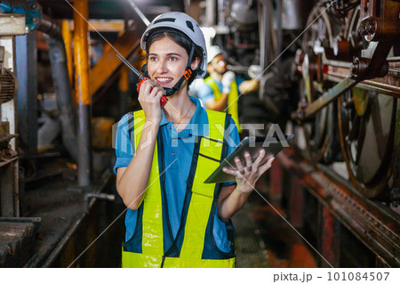 Beautiful woman engineer in factory talking on the radio wearing white helmet and work clothes and hand holding tablet at workplace Beautiful woman engineer in factory talking on the radio wearing white helmet and work clothes and hand holding tablet at workplace 101084507