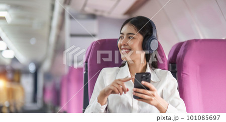 Cheerful female passenger in headphones for noise cancellation watching online movie during intercontinental flight in cabin of aircraft, happy young woman using wifi connection on board 101085697