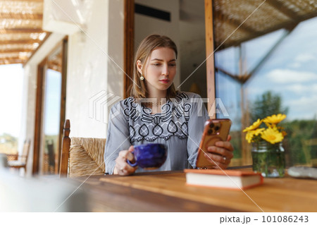 Young woman in ethnic outfit sitting at table with phone, working, having mug of morning coffee at home. 101086243