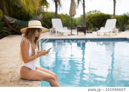 Girl in swimsuit and hat using mobile phone sitting and relaxing near swimming pool. Girl in swimsuit and hat using mobile phone sitting and relaxing near swimming pool. 101086607