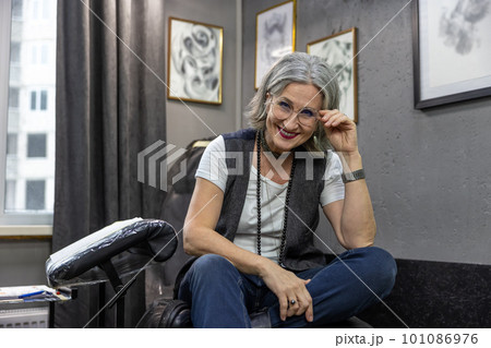 Grey-haired woman sitting on couch in the tattoo salon Grey-haired woman sitting on couch in the tattoo salon 101086976