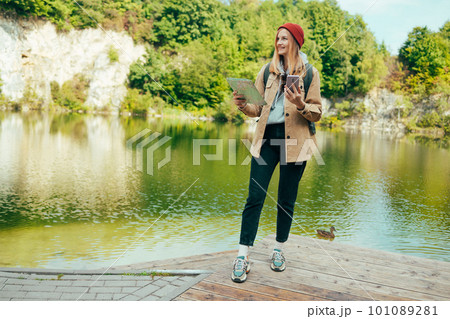 Happy woman tourist with backpack and red hat holding paper map walking in mountains with lake view background. Travel and wanderlust concept. Amazing chill moment.  101089281