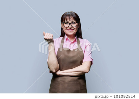 Positive smiling mature woman in apron on gray studio background Positive smiling mature woman in apron on gray studio background 101092814