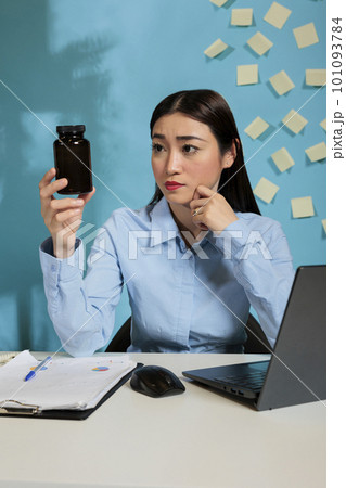 Professional asian startup employee with headache looking at bottle of pills analyzing the components of contains. Woman at office desk looking doubtfully at a container of tablets. Professional asian startup employee with headache looking at bottle of pills analyzing the components of contains. Woman at office desk looking doubtfully at a container of tablets. 101093784