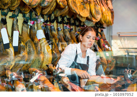 Young saleswoman in black apron slicing Iberian jamon at counter in butcher shop Young saleswoman in black apron slicing Iberian jamon at counter in butcher shop 101097052