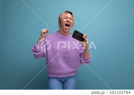 energetic blond young woman consultant in purple hoodie holding smartphone with mockup and singing 101103794