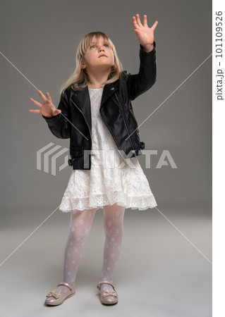 Little blonde girl in a white dress and black leather jacket is posing standing over a gray background. Little blonde girl in a white dress and black leather jacket is posing standing over a gray background. 101109526