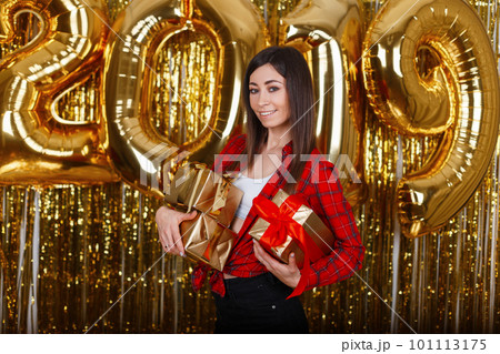 Young joyful woman in a stylish red shirt and jeans holds in her hands shiny gift boxes on the background of golden balls of 2019. Christmas positive mood. Happy cheerful girl. Party is on. Young joyful woman in a stylish red shirt and jeans holds in her hands shiny gift boxes on the background of golden balls of 2019. Christmas positive mood. Happy cheerful girl. Party is on. 101113175