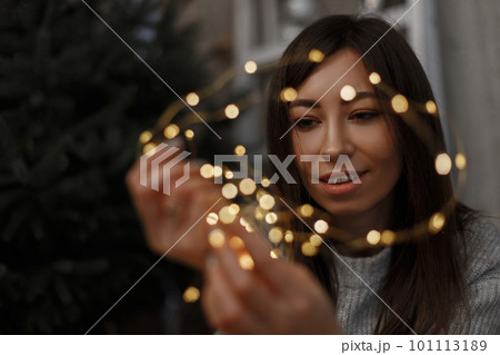 Young happy beautiful girl in a gray fashionable sweater with Christmas lights in her hands. Friendly girl smiling. Warm New Year mood. 101113189