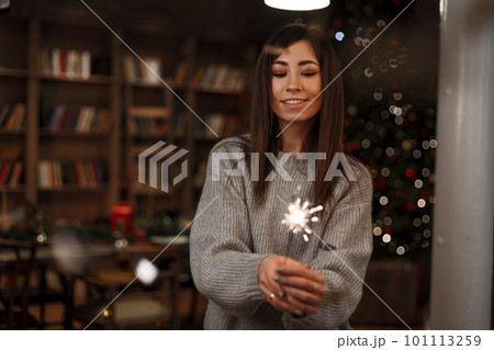 Happy young woman in a knitted sweater is standing with a sparkler in her hand and is looking at him against the background of an vintage bookcase in a festive room. Joyful girl. Happy young woman in a knitted sweater is standing with a sparkler in her hand and is looking at him against the background of an vintage bookcase in a festive room. Joyful girl. 101113259