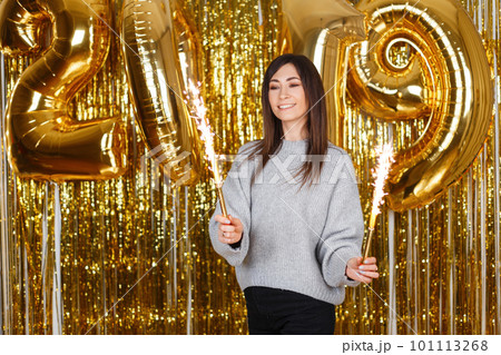 Positive young woman in a vintage knitted sweater with fireworks on the background of festive gold garlands and with New Year's air balloons 2019. Girl with a beautiful smile celebrates the New Year. 101113268