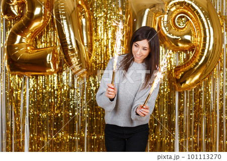 Happy young woman in a gray knitted sweater with fireworks on the background of golden garlands and New Year's balloons 2019. Girl with a beautiful smile is fun at the New Year's party. Happy young woman in a gray knitted sweater with fireworks on the background of golden garlands and New Year's balloons 2019. Girl with a beautiful smile is fun at the New Year's party. 101113270