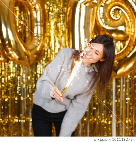 Young pretty woman in knitted sweater enjoy with fireworks in hands near the background of golden balloons 2019 and garlands in a festive studio. Girl is fun at the New Year's party. 101113275