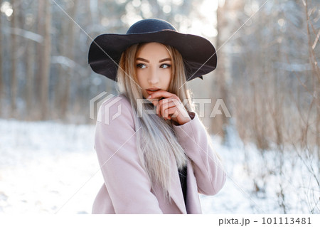 Amazing glamorous pretty beautiful young woman in a luxurious black hat in a stylish pink warm coat posing on a sunny day in a winter park. Sexy fashionable girl on a walk. Amazing glamorous pretty beautiful young woman in a luxurious black hat in a stylish pink warm coat posing on a sunny day in a winter park. Sexy fashionable girl on a walk. 101113481