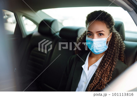 Woman in a sterile mask in taxi car on a back seat looking out of window checking her cell phone. 101113816