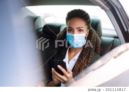 Woman in a sterile mask in taxi car on a back seat looking out of window checking her cell phone. 101113817