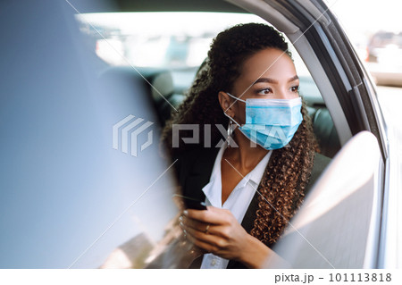 Woman in a sterile mask in taxi car on a back seat looking out of window checking her cell phone. 101113818
