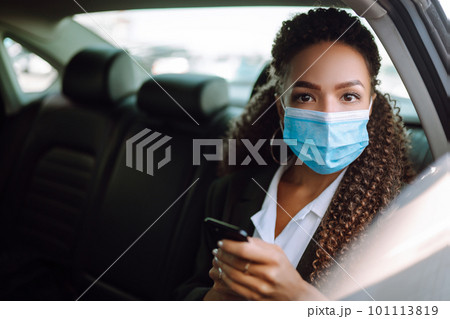 Woman in a sterile mask in taxi car on a back seat looking out of window checking her cell phone. 101113819