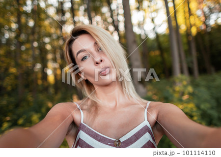 Cute funny young woman blonde in a vintage pink striped dress resting in the forest on a sunny day and taking pictures of herself on the phone. Happy girl makes selfie. 101114110