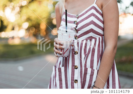 Glamorous young woman in a stylish striped pink dress poses in the park and holds in hands a sweet milkshake. Modern fashionable girl with a cool tasty dessert. Hot summer sunny day. 101114179