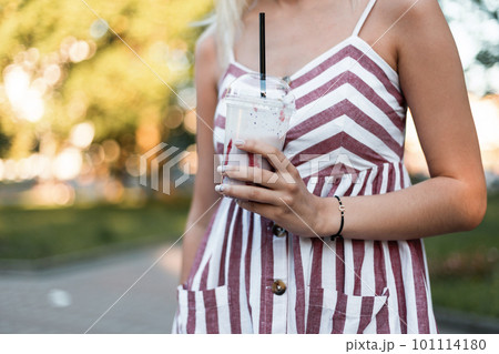 Close-up of a young woman in a summer pink striped dress with a milkshake in a sunny day outdoors in the park. Stylish girl enjoys a walk and a sweet drink outdoors in the park. 101114180