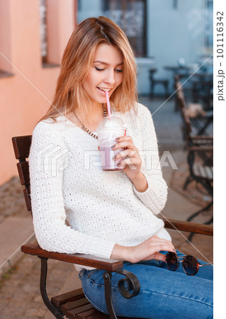 Happy charming young woman in trendy summer clothes is sitting in a vintage street cafe and drinking a sweet cocktail. Cheerful joyful stylish girl. 101116342