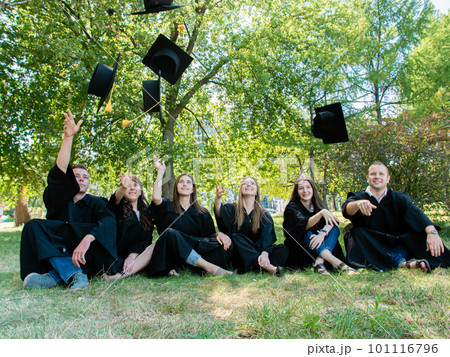 Happy student friends in mantle sit on the grass in the university park. Graduates throw their academic cap up Happy student friends in mantle sit on the grass in the university park. Graduates throw their academic cap up 101116796