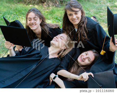 Several young smiling female students in black graduation gowns lie on the grass in the university park 101116860