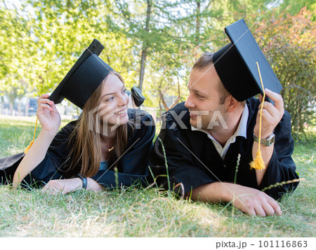 Happy guy and girl look at each other lying on the lawn in a black graduate gown and an academic cap of the university Happy guy and girl look at each other lying on the lawn in a black graduate gown and an academic cap of the university 101116863