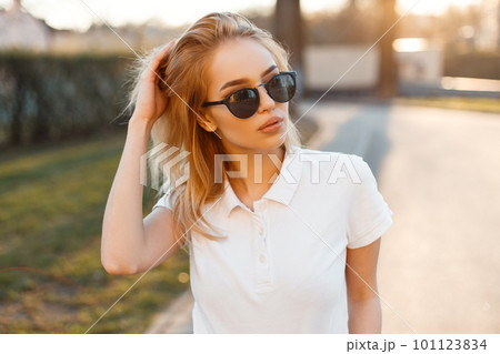 Modern stylish young hipster woman in trendy white polo t-shirt in black sunglasses stand and posing on the street against the background of the sun. American beautiful girl. Modern stylish young hipster woman in trendy white polo t-shirt in black sunglasses stand and posing on the street against the background of the sun. American beautiful girl. 101123834