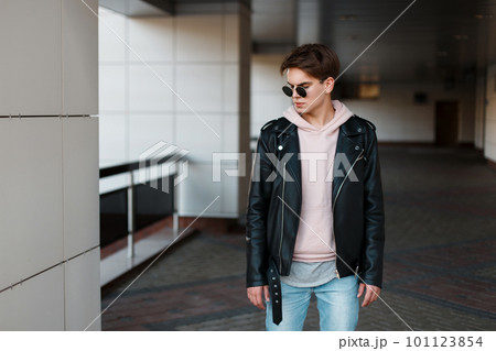 Fashionable young hipster man in black sunglasses in stylish black jacket in pink sweatshirt in trendy jeans in a gray t-shirt is standing in the room. Serious cool guy with a beautiful hairstyle Fashionable young hipster man in black sunglasses in stylish black jacket in pink sweatshirt in trendy jeans in a gray t-shirt is standing in the room. Serious cool guy with a beautiful hairstyle 101123854