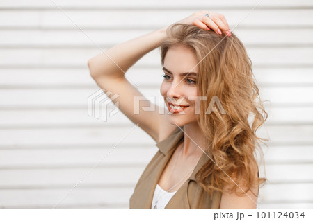 Cheerful charming young woman with a wonderful smile with curly blond hair in a stylish waistcoat posing near a white wooden vintage wall on a warm summer day. Happy positive stylish girl. 101124034