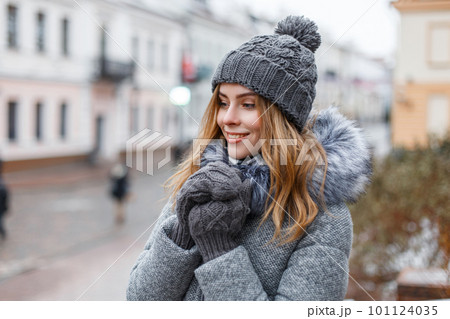Happy attractive young woman in a winter knitted hat in a trendy gray coat with fur in knitted warm mittens with a beautiful smile walks around the city on a winter day. Cheerful girl enjoy a walk. Happy attractive young woman in a winter knitted hat in a trendy gray coat with fur in knitted warm mittens with a beautiful smile walks around the city on a winter day. Cheerful girl enjoy a walk. 101124035
