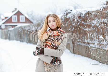 Young woman in a vintage gray coat in a woolen stylish scarf in warm woolen mittens stands in the winter afternoon on the background of a rustic wooden house. Lovely girl walks through the countryside 101124037