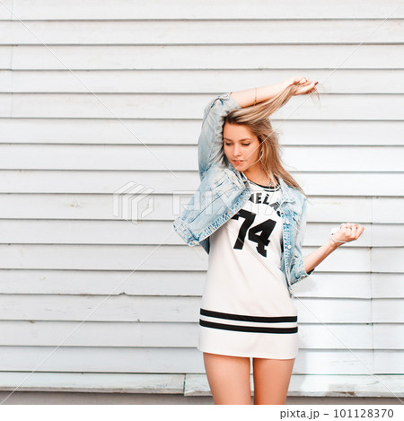 Cheerful attractive young woman in a sporty white summer dress with a black pattern in a denim jacket is dancing near a wooden vintage building on a warm summer day. Happy girl enjoys the weekend. 101128370