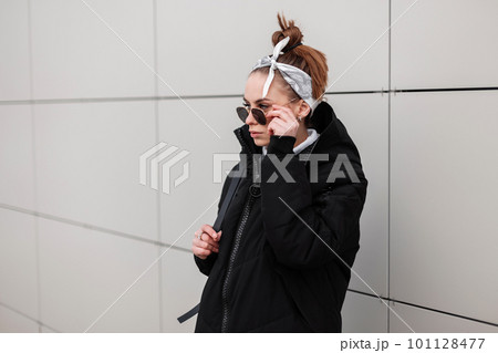 Young woman hipster in a stylish long jacket with a leather backpack with a stylish hairstyle in a trendy bandana in black sunglasses near a white wall in the outdoors. American girl. Modern fashion. 101128477