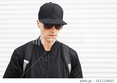 Portrait of a fashionable man hipster in a black cap in stylish black sunglasses in a trendy jacket with a black backpack on his shoulders on a white background. Serious stylish American guy. Portrait of a fashionable man hipster in a black cap in stylish black sunglasses in a trendy jacket with a black backpack on his shoulders on a white background. Serious stylish American guy. 101128683