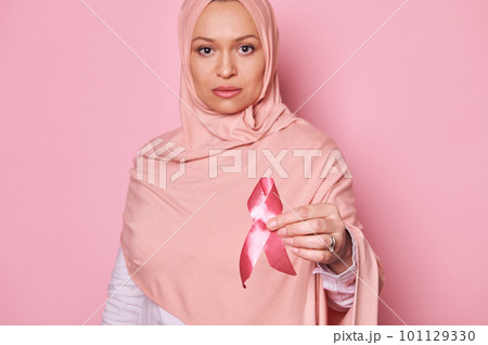 Selective focus on Breast Cancer Awareness Pink Ribbon, in hands of blurred Arab Muslim woman in pink hijab, showing her support for cancer survivors and patients, isolated background. 1 st October 101129330