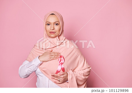 Confident Arab Muslim woman in hijab, wearing pink satin awareness ribbon, emblem of fighting Breast Cancer and supporting patients and survivors, looking confidently at camera on isolated background Confident Arab Muslim woman in hijab, wearing pink satin awareness ribbon, emblem of fighting Breast Cancer and supporting patients and survivors, looking confidently at camera on isolated background 101129896