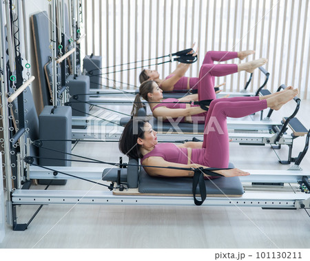 Three Asian women in pink sportswear doing pilates exercises with a reformer bed. Three Asian women in pink sportswear doing pilates exercises with a reformer bed. 101130211