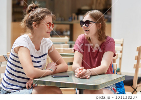 Joyful female companions in shades, have friendly talk in coffee shop while wait for order, have fun together, recreat during summer time, gossip about other people. Friends and leisure concept 101138237
