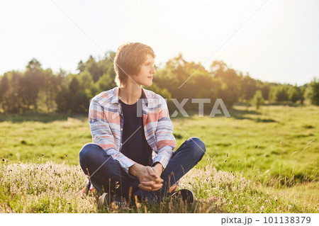 Serious thoughtful boy in casual clothes sitting crossed legs on green grass looking aside with dreamy expression admiring sunlight or beautiful nature. Young male sitting outdoors on meadow 101138379