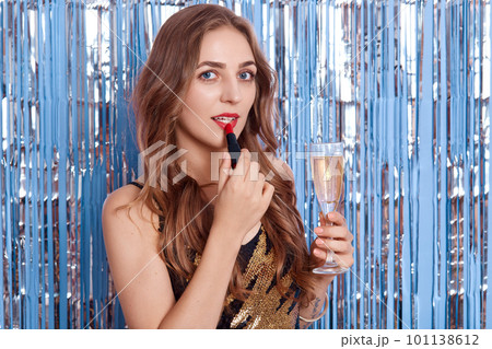Cheerful attractive young woman with bright makeup, holding glass of wine or champagne, lady applies pomade, girl posing isolated over blue background decorated with glitter tinsel. 101138612