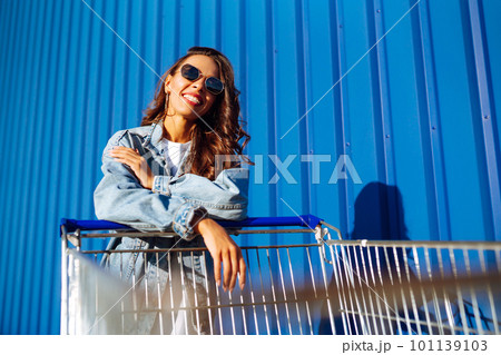 Young woman with phone sitting shopping cart having fun near color wall. Blogging. Shopaholic. 101139103