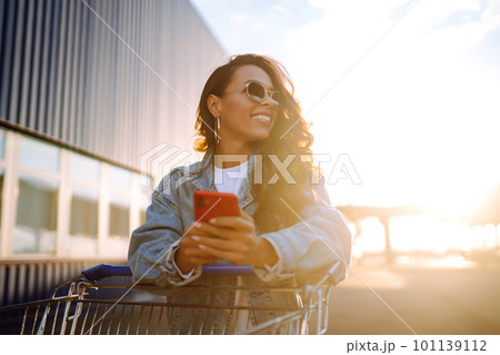 Young woman with phone sitting shopping cart having fun near color wall. Blogging. Young woman with phone sitting shopping cart having fun near color wall. Blogging. 101139112