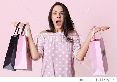Indoor shot of beautiful fashionable Caucasian woman holding shopping bags in both hands, has shocked facial expression, posing with opened mouth isolated over pink background. Shopping concept. 101139217
