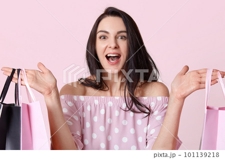 Headshot of pleased dark haired young woman holds shopping bags, looks joyfully, rejoices good day, makes successful purchases, models over light pink background. People and purchasing concept 101139218